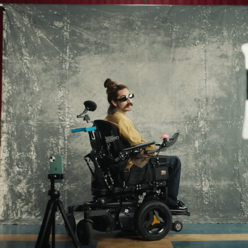 A fashionable man in a power wheelchair uses Apple Voice Control to tell his iPhone when to snap photos.